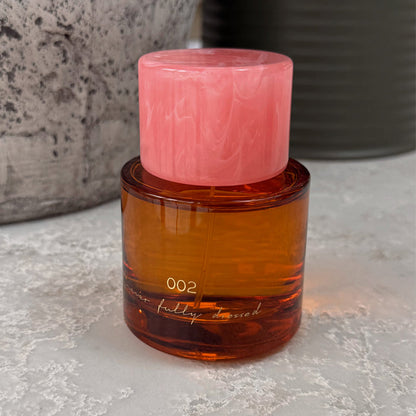 NFD Eau De Parfum 002 bottle in amber glass with a pink marbled cap, set on a textured grey stone surface.