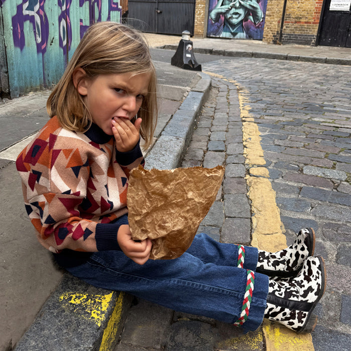 NFD Kids Aspen Jumper with an orange and navy geometric pattern, worn by a child sitting on a London curb.