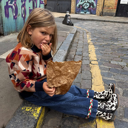 NFD Kids Aspen Jumper with an orange and navy geometric pattern, worn by a child sitting on a London curb.
