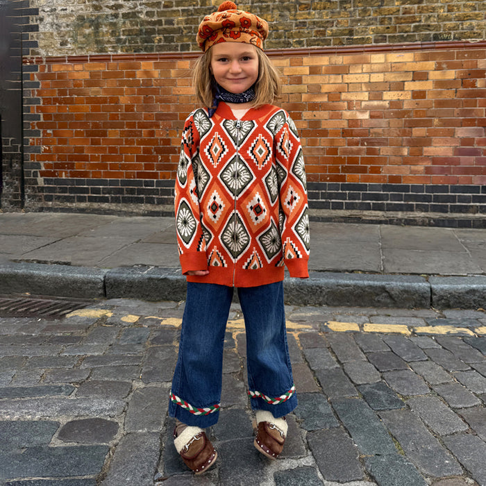 Rust orange NFD Kids Diamond Bomber with an olive and red knitted diamond pattern, worn by a child on a cobbled street.