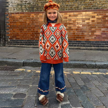 Rust orange NFD Kids Diamond Bomber with an olive and red knitted diamond pattern, worn by a child on a cobbled street.