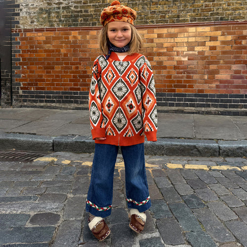 Rust orange NFD Kids Diamond Bomber with an olive and red knitted diamond pattern, worn by a child on a cobbled street.