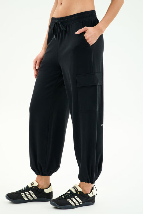A person wears the SPLITS59 Nico Airweight Cargo Pant in black, paired with black sneakers with white stripes and a cropped black top, standing with one hand in a pocket for effortless comfort.