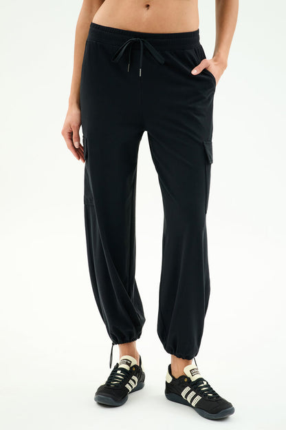 A person wears SPLITS59 Nico Airweight Cargo Pant in black for lightweight comfort, paired with black athletic shoes, standing against a plain light background.