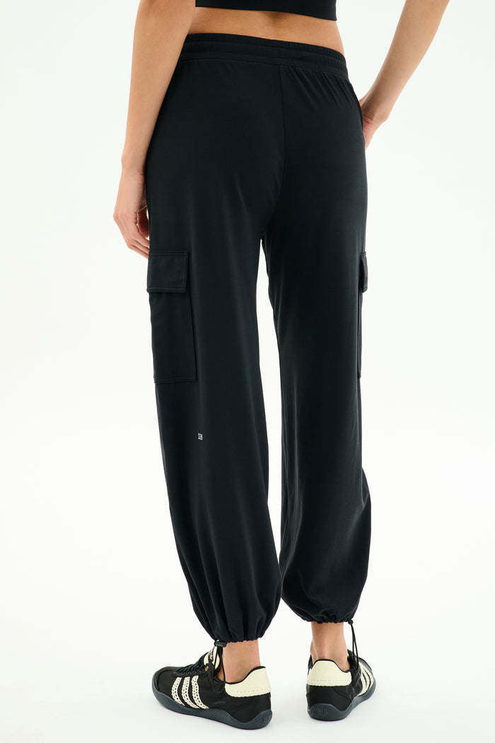 A person wears the SPLITS59 Nico Airweight Cargo Pant in black, known for its lightweight comfort, paired with black athletic shoes, standing against a plain white background.