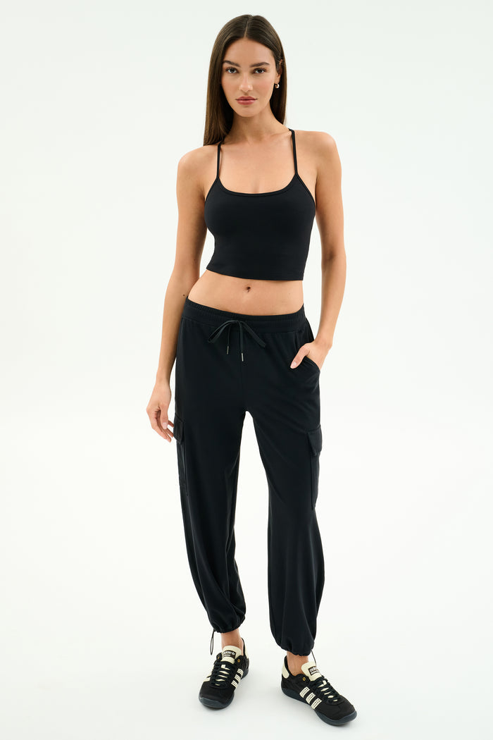 A woman stands against a white background wearing a black tank top, black SPLITS59 Nico Airweight Cargo Pant for lightweight comfort, and black sneakers with white stripes.