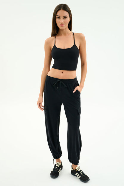 A woman stands against a white background wearing a black tank top, black SPLITS59 Nico Airweight Cargo Pant for lightweight comfort, and black sneakers with white stripes.