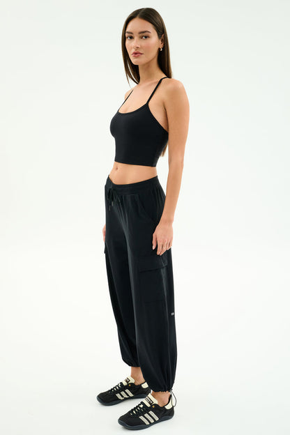 A woman poses against a plain background in the SPLITS59 Nico Airweight Cargo Pant in black, paired with a black crop top and black sneakers with white stripes.