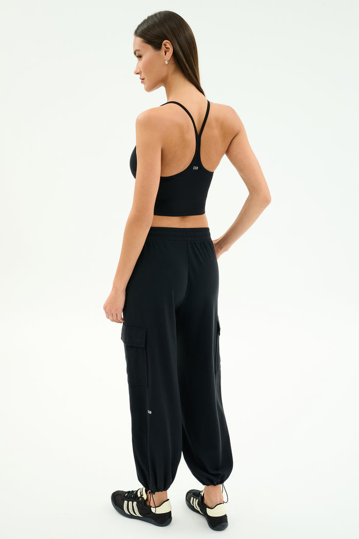 A woman faces away, wearing a black sports bra, SPLITS59 Nico Airweight Cargo Pant in black for lightweight comfort, and black Adidas sneakers against a plain white background.