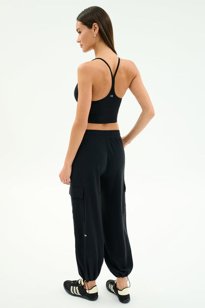 A woman faces away, wearing a black sports bra, SPLITS59 Nico Airweight Cargo Pant in black for lightweight comfort, and black Adidas sneakers against a plain white background.