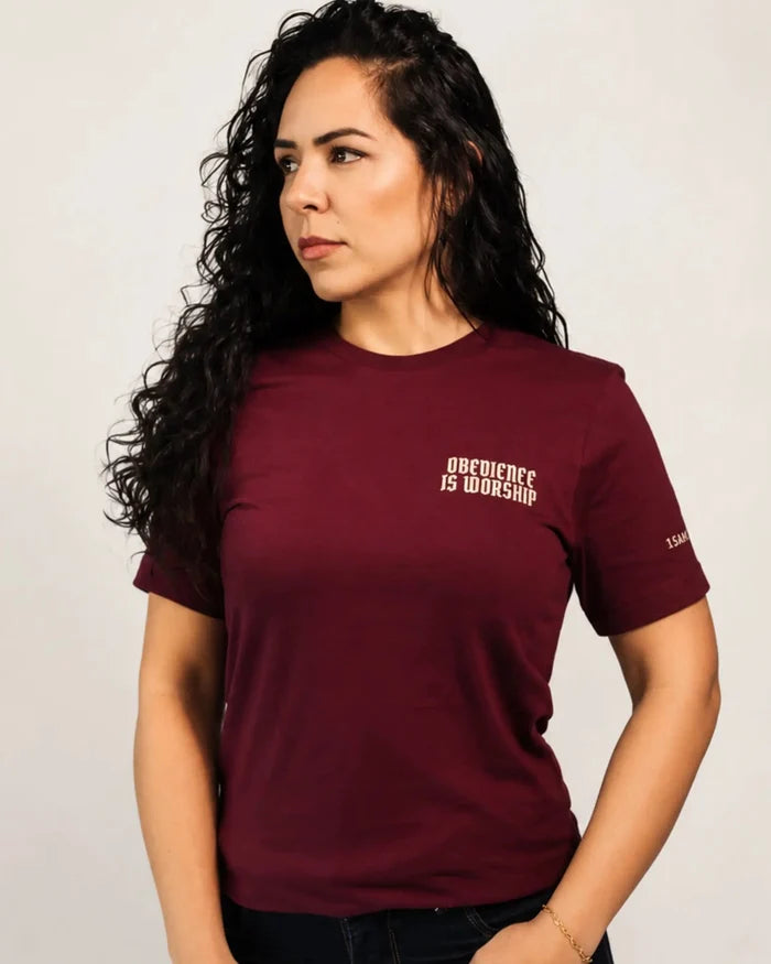 A woman with long, curly dark hair wearing a maroon short-sleeve t-shirt with white text on the left chest that reads 'OBEYIENCE IS WORSHIP' and a small white logo on the right sleeve. She is posing against a light gray studio background.