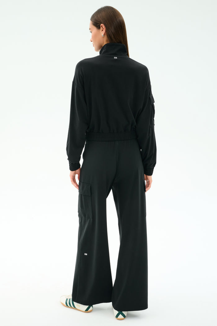 A person faces away, wearing the SPLITS59 Peaches Airweight Cargo Jacket in black stretch fabric and wide-leg pants with white text, plus white slide sandals with green stripes, against a plain light background.