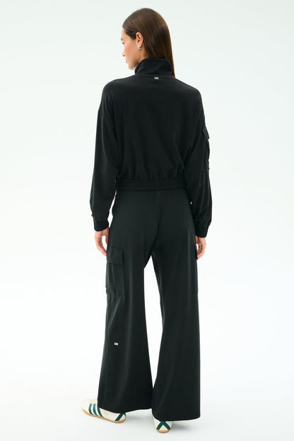 A person faces away, wearing the SPLITS59 Peaches Airweight Cargo Jacket in black stretch fabric and wide-leg pants with white text, plus white slide sandals with green stripes, against a plain light background.