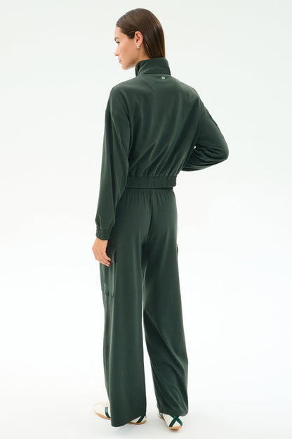 A woman stands with her back to the camera, wearing the SPLITS59 Peaches Airweight Cargo Jacket in Military, featuring soft nylon spandex, a high collar, and wide-leg pants, paired with white sneakers on a plain light background.