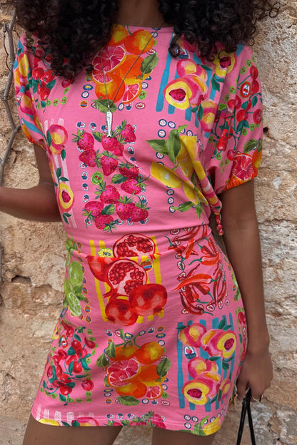 Pink Fruit Stamps Mini Dress with a fruit motif and t-shirt silhouette, styled against a textured stone wall.