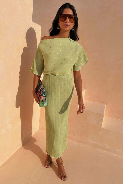 Pistachio Tilly Dress in green plisse with gold flecks and a lettuce hem, styled off-the-shoulder on stairs.