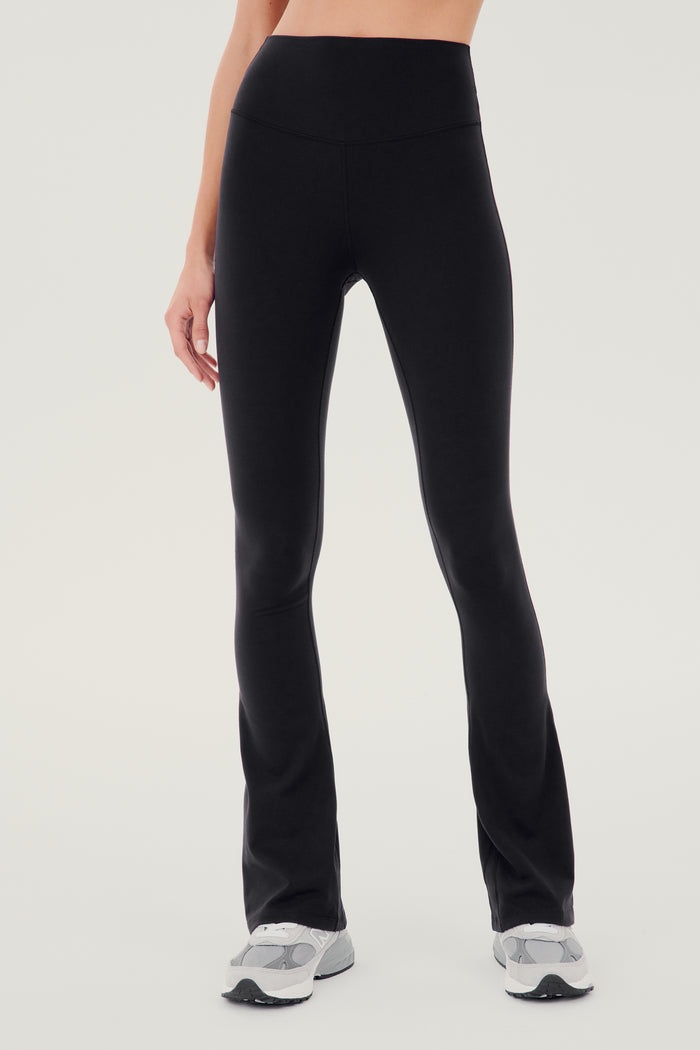 Front view of woman wearing black high waist below ankle length legging with wide flared bottoms. Paired with white and gray shoes.
