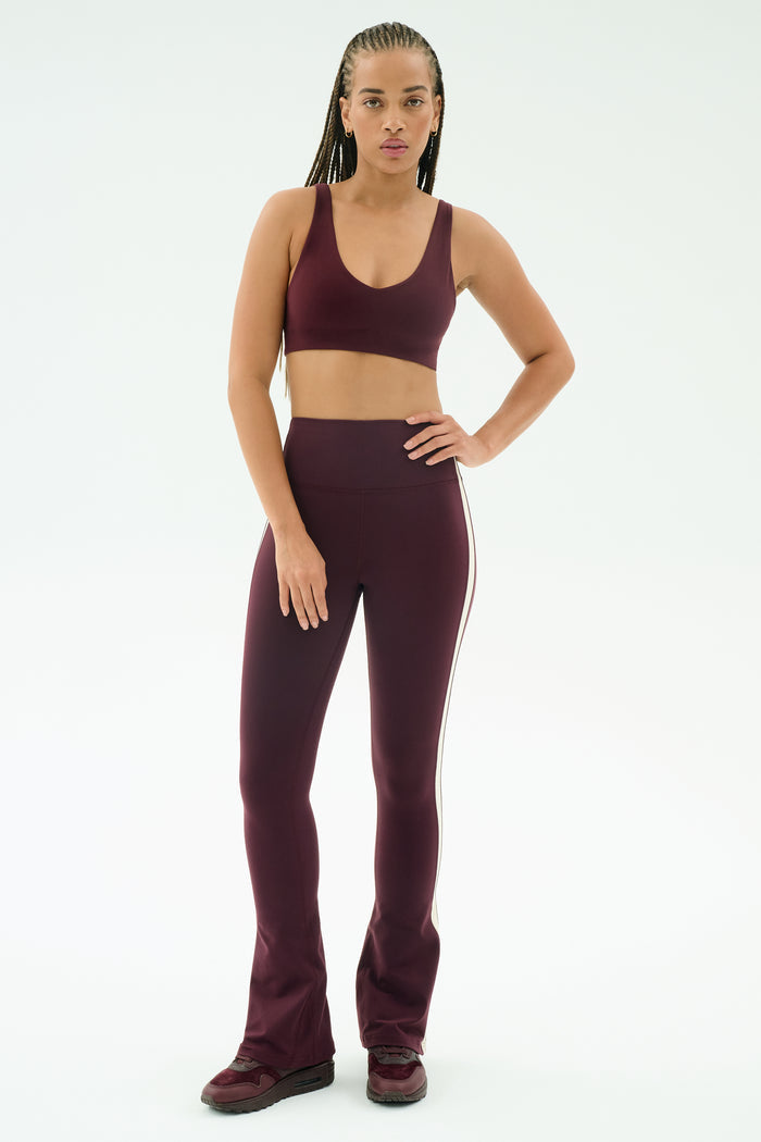 A person stands facing forward, wearing SPLITS59's Raquel High Waist Flared Legging in Dark Cherry/Creme with a matching sports bra and sneakers, set against a plain white background.