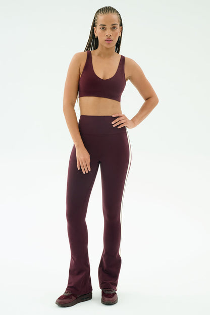 A person stands facing forward, wearing SPLITS59's Raquel High Waist Flared Legging in Dark Cherry/Creme with a matching sports bra and sneakers, set against a plain white background.