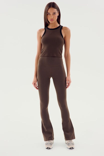 A woman faces forward, wearing SPLITS59's Raquel High Waist Flared Legging in Dark Chocolate with a sleeveless brown top and white sneakers, against a plain white background.