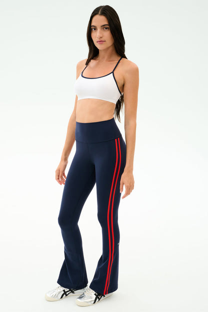 A woman wears the SPLITS59 Raquel High Waist Flared Legging in Indigo/Lotus Red, paired with a white sports bra and white sneakers, standing against a plain light background. The leggings offer 4-way stretch for comfort and performance.