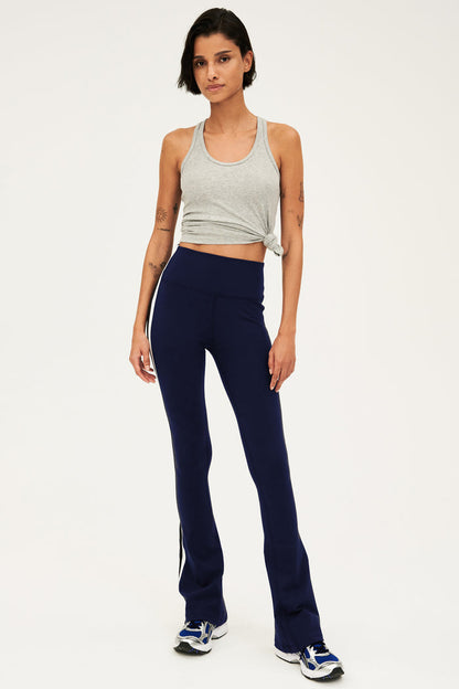 Full front view of woman with dark short hair wearing dark blue  high waist below ankle length legging with wide flared bottoms and white and black side stripes on both legs. Also wearing a dark gray racerback tank top with right knot on the bottom. Paired with gray,black and bright blue shoes.