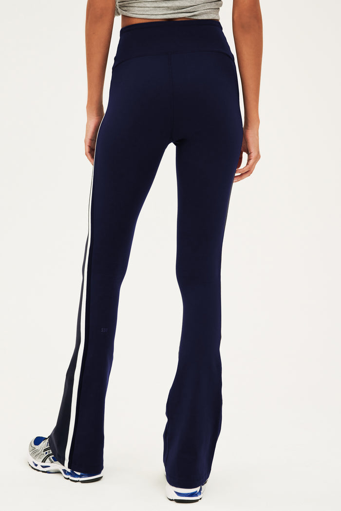 Back view of woman wearing dark blue high waist below ankle length legging with wide flared bottoms with gray S59 logo on back of left calf  and white and side black stripes on both legs. Paired with gray,black and bright blue shoes.
