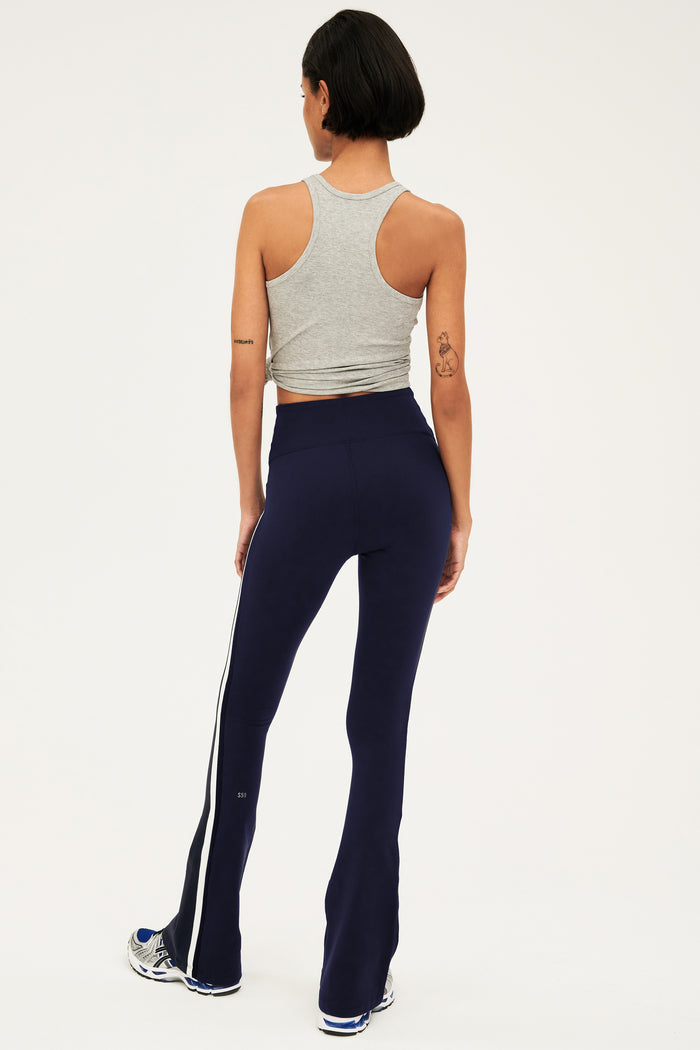 Full back view of woman with dark short hair wearing dark blue  high waist below ankle length legging with wide flared bottoms with gray S59 logo on back of left calf and white and side  black stripes on both legs. Also wearing a dark gray racerback tank top. Paired with gray,black and bright blue shoes.