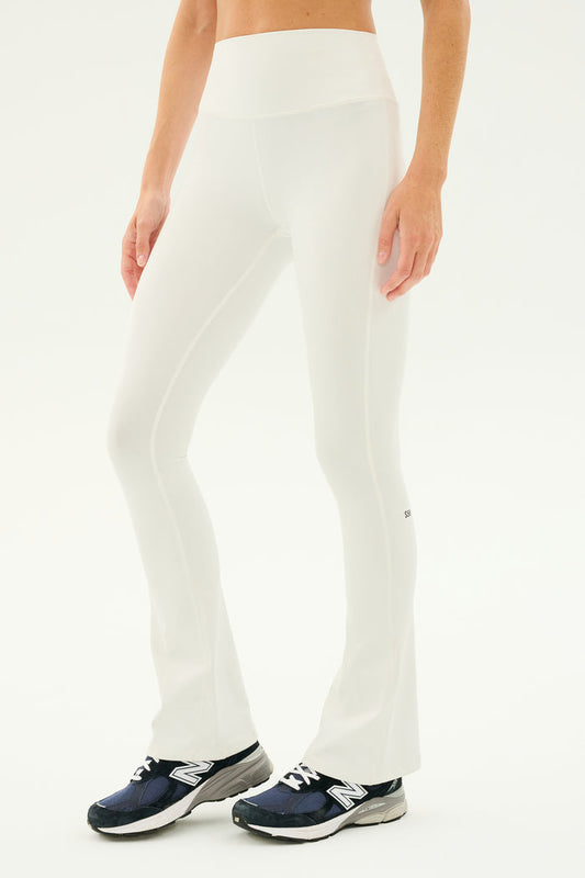Raquel stands against a plain background, showcasing her SPLITS59 Raquel High Waist Flared Legging in white, made from 4-way stretch Supplex fabric, paired with black athletic shoes.