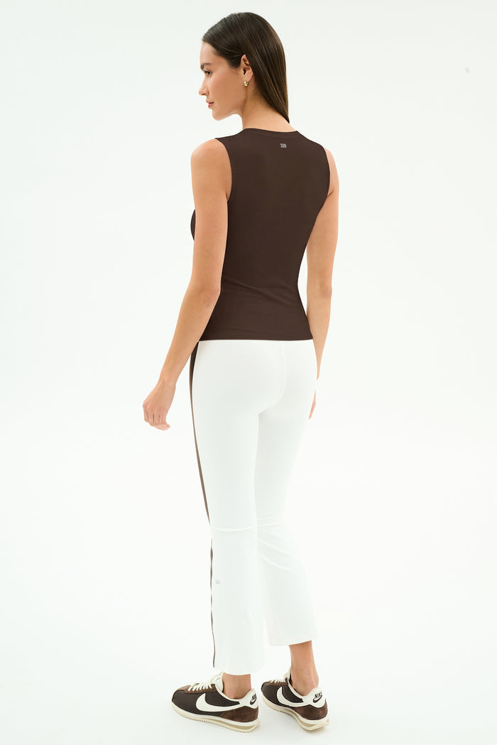 A woman stands facing away, wearing a sleeveless dark brown top, SPLITS59 Raquel High Waist Supplex Crop leggings in White/Dark Chocolate, and black Nike sneakers against a plain white background.