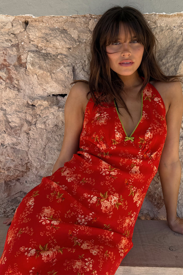 Red Floral Halter Dress with green trim and bow detailing, worn by a model against a rustic stone wall background.