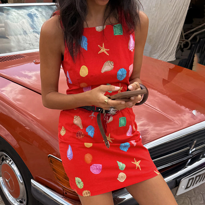 Red Shell Ioana Mini Dress in a cotton-linen blend with a multicoloured shell print, leaning against a vintage car.