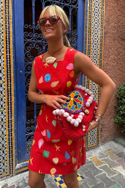 Red Shell Ioana Mini Dress in cotton-linen with a multicoloured shell print, red sunglasses and a crochet bag.