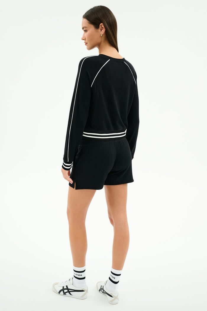 Someone stands facing away, wearing the SPLITS59 Reena Fleece Sweatshort in black, a black long-sleeve top with white trim, white socks, and white athletic sneakers against a plain light background.