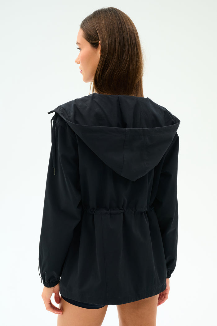 A woman faces away, wearing the SPLITS59 Reggie Water Repellent Jacket in black/white, featuring a cinched waist and sleek nylon fabric.
