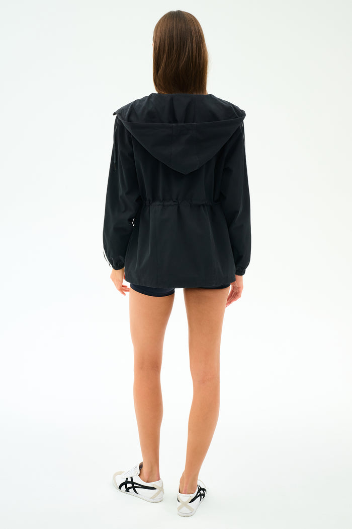 A woman faces away, wearing the SPLITS59 Reggie Water Repellent Jacket in black/white, along with black spandex shorts and white sneakers, against a plain white background.