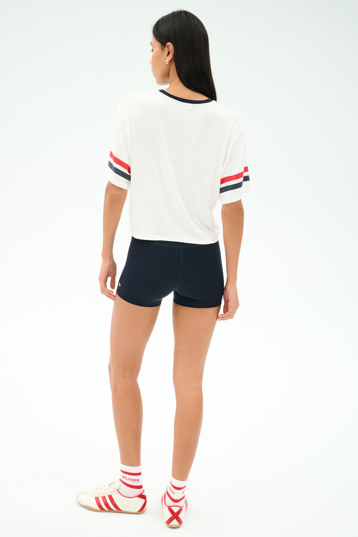 A person stands on a white background wearing the SPLITS59 Rex Varsity Jersey V-Neck in White/59, black shorts, white socks with red stripes, and white sneakers with red details.