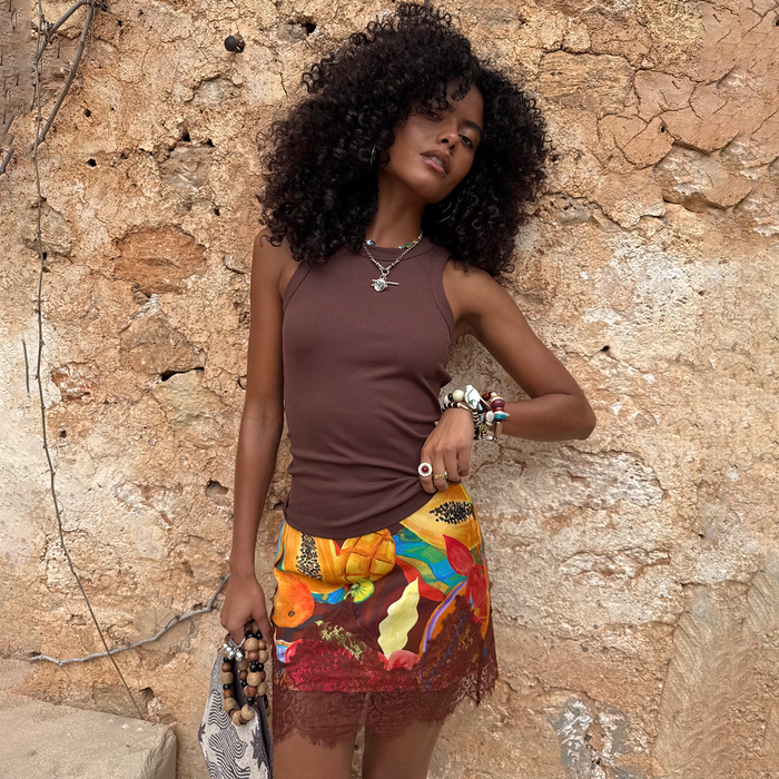 Rio Mini Maeva Skirt in a tropical fruit print with brown lace trim, styled on a model against a stone wall.