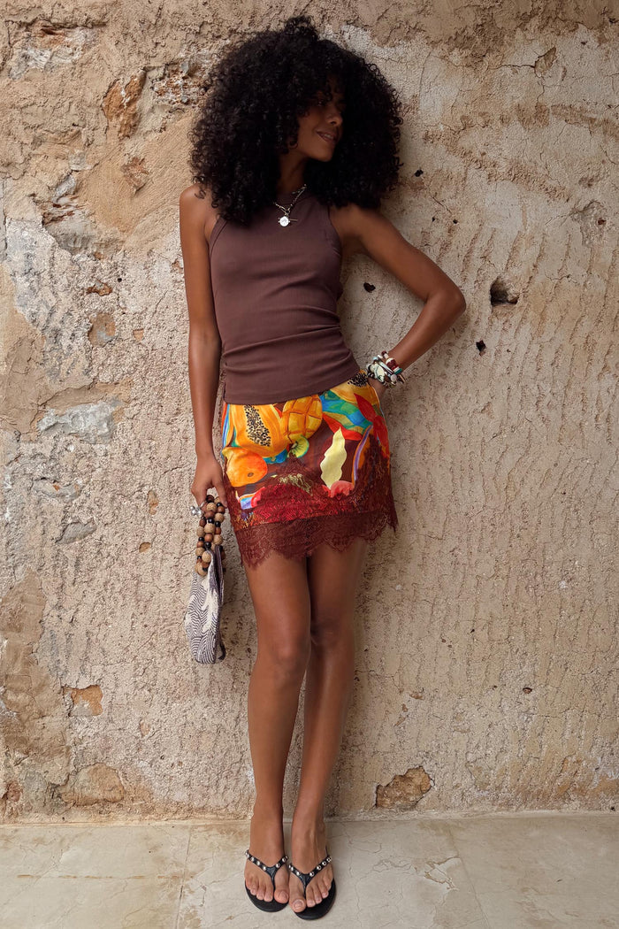 Rio Mini Maeva Skirt in a tropical fruit print with brown lace trim, styled with a brown vest for a summer vacation look.