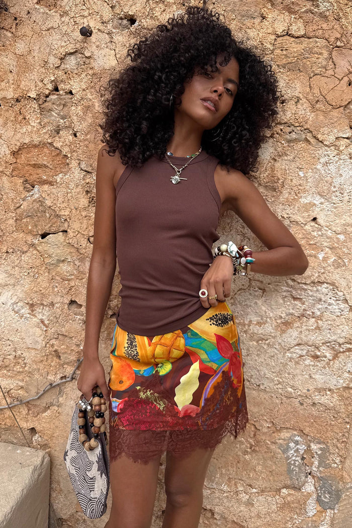 Rio Mini Maeva Skirt featuring a tropical fruit print and brown lace trim, worn by a model against a stone wall.