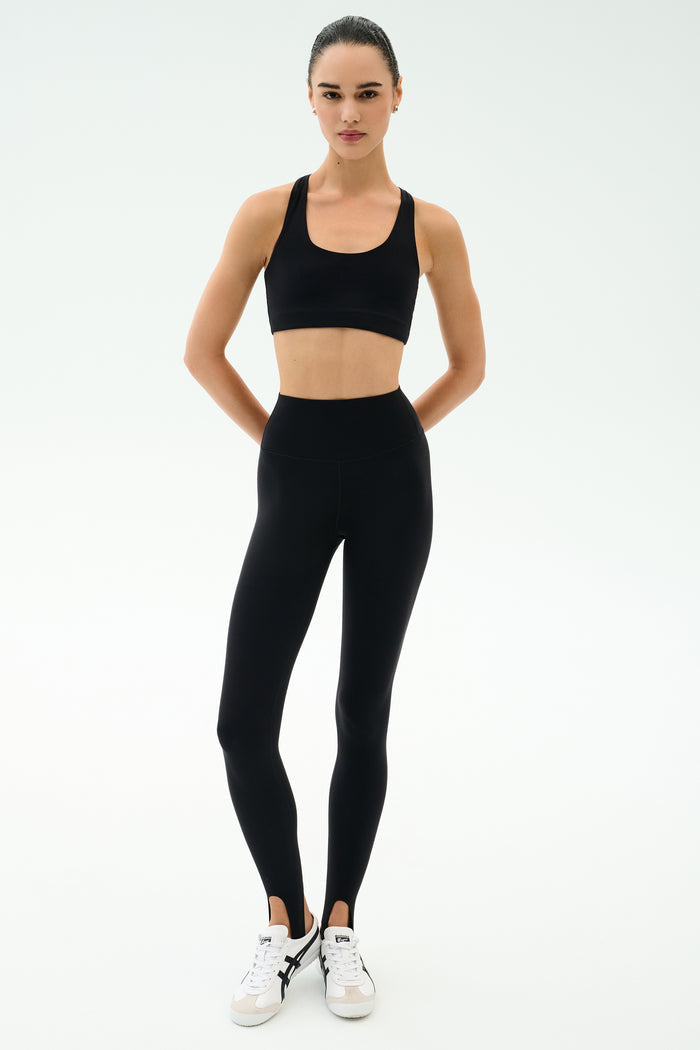 A person stands facing forward, wearing SPLITS59 River High Waist Airweight Stirrup leggings in black and a black sports bra, paired with white sneakers against a plain light background.