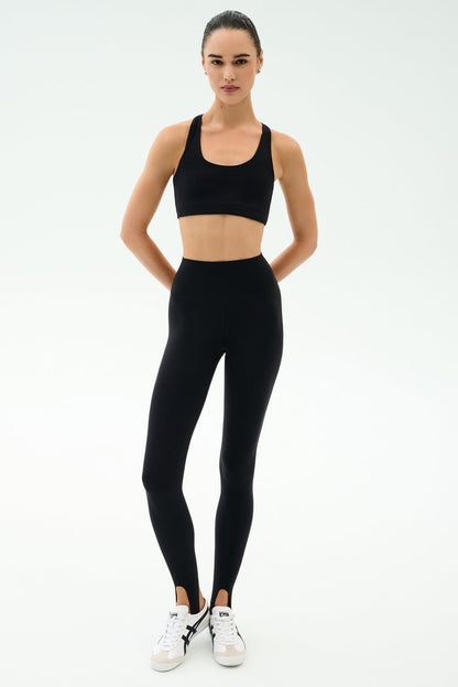 A person stands facing forward, wearing SPLITS59 River High Waist Airweight Stirrup leggings in black and a black sports bra, paired with white sneakers against a plain light background.