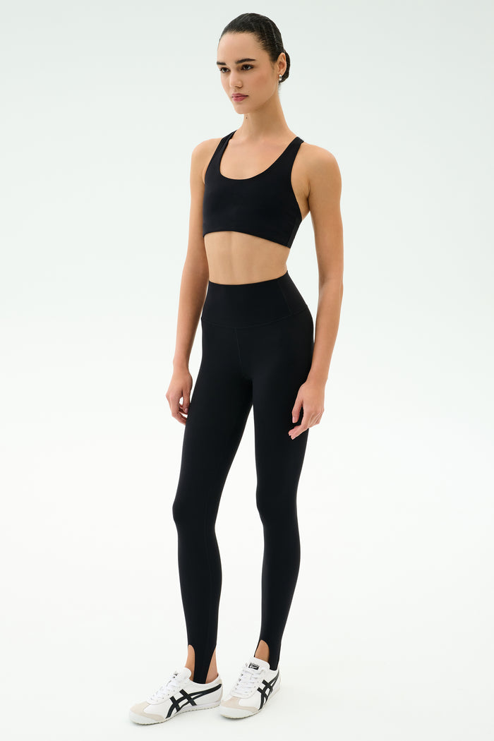 A person stands facing forward wearing the SPLITS59 River High Waist Airweight Stirrup leggings in black with white sneakers, set against a plain light background.