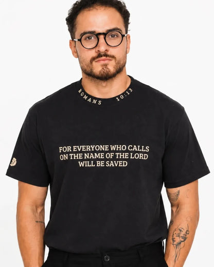 A man wearing a black crewneck t-shirt with gold text that reads 'FOR EVERYONE WHO CALLS ON THE NAME OF THE LORD WILL BE SAVED' and 'ROMANS 10:13' printed around the collar, set against a plain white background.