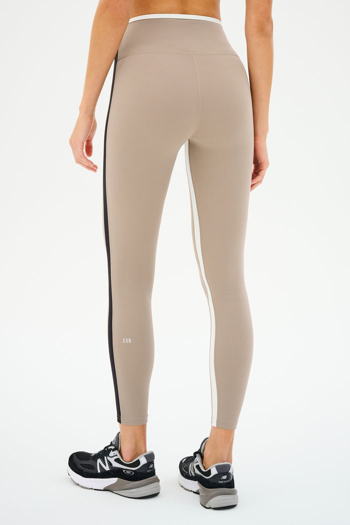A person, stylishly photographed from the back, pairs sleek black sneakers with SPLITS59's Sam High Waist Rigor 7/8 leggings in Latte/Black featuring side stripes.