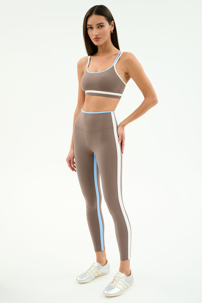 A woman stands in SPLITS59’s Sam High Waist Rigor 7/8 leggings in Lentil/White, featuring compressive fabric and blue and white stripes, paired with a taupe sports bra and white sneakers against a light background.