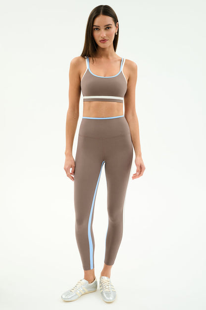 A woman faces forward in SPLITS59’s Sam High Waist Rigor 7/8 leggings in Lentil/White with a matching taupe sports bra and silver sneakers, standing against a plain white background.