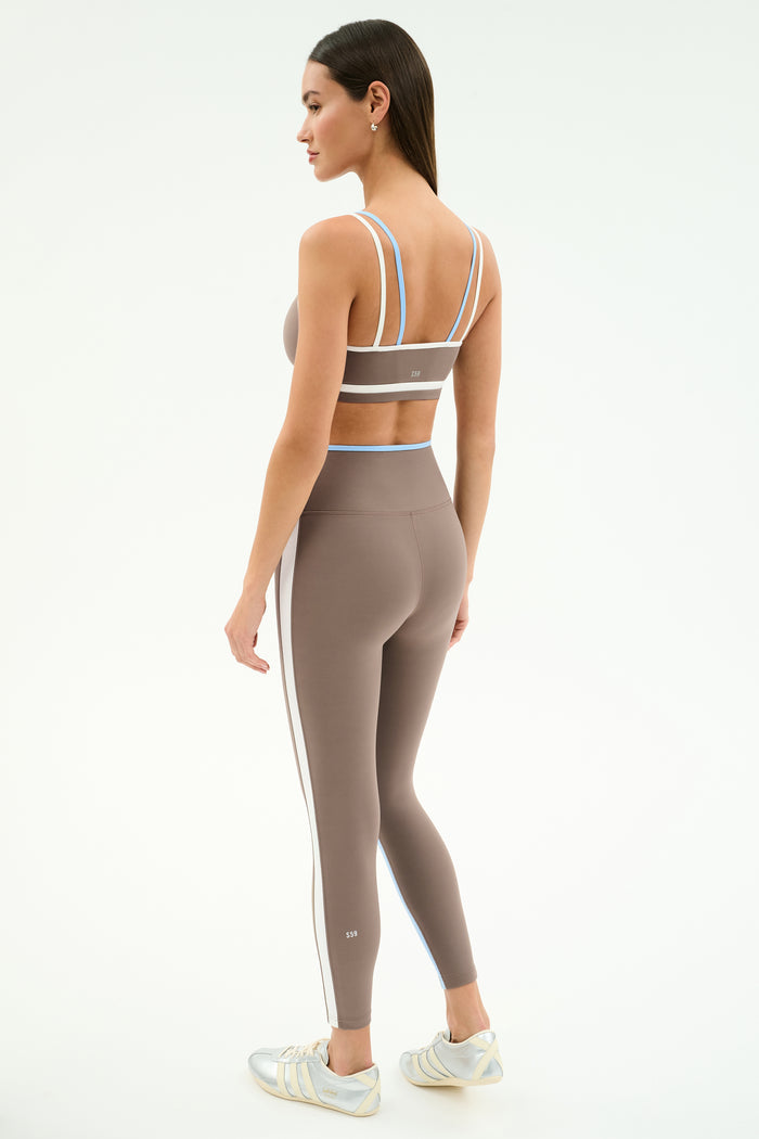A woman stands sideways in SPLITS59's Sam High Waist Rigor 7/8 leggings in Lentil/White with matching taupe sports bra, featuring compressive fabric and white stripes, paired with light-colored athletic shoes against a plain background.