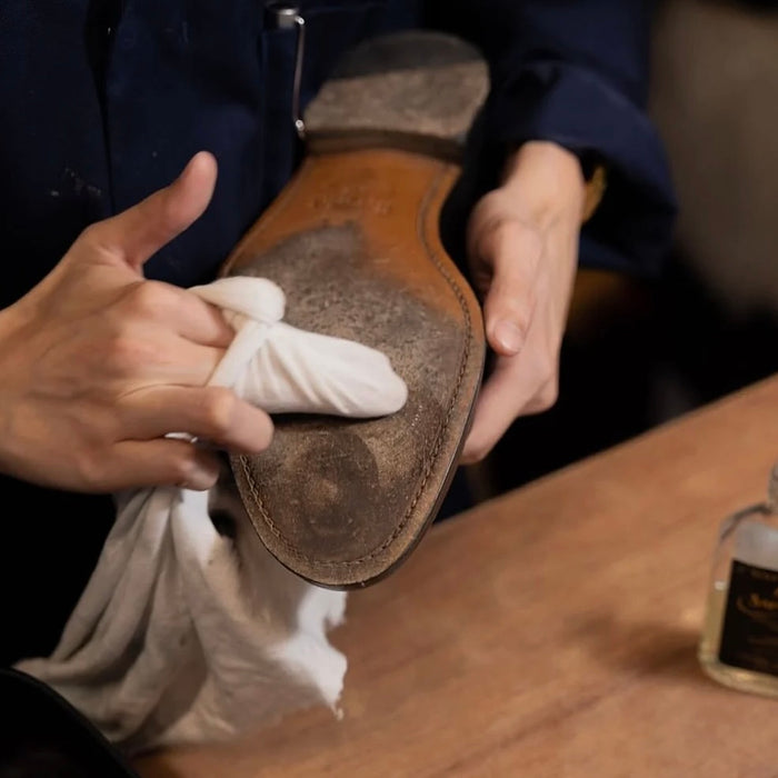 Person cleaning a leather shoe with a white cloth on a wooden surface.