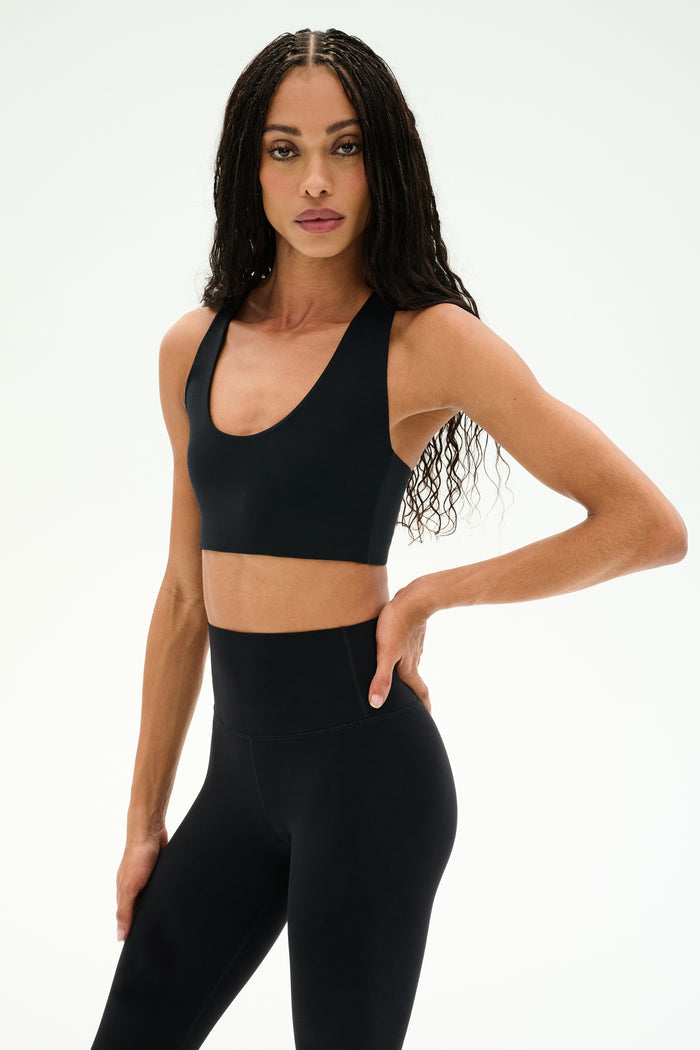 A woman with long braided hair, dressed in the SPLITS59 Sara Airweight Bra in black and high-waisted leggings, stands against a plain light background facing forward with one hand on her hip—perfectly outfitted for pilates or hot yoga.